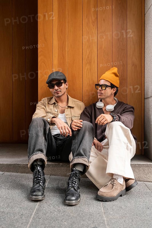 Two fashionable gay men sit on steps against a wooden backdrop. Both exhibit modern style with hats and sunglasses, emphasizing a relaxed urban vibe