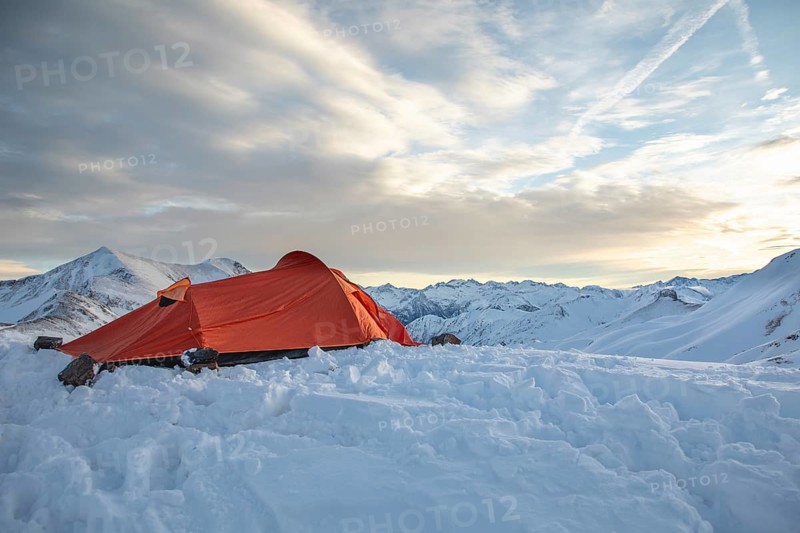 A vibrant orange tent sits atop a snow-covered peak, surrounded by majestic mountain views under a clear sky, ideal for ski touring or alpine adventure enthusiasts