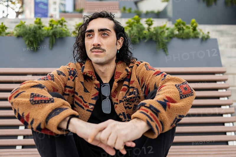 A man with curly hair and a mustache sits on an outdoor bench wearing a patterned jacket and sunglasses The urban background adds to his relaxed, stylish vibe