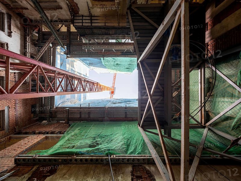 Looking up inside a building under construction, steel beams, scaffolding, and a towering crane frame the sky. Green safety netting covers areas for protection