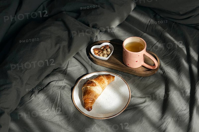 A golden homemade croissant rests on a white plate, accompanied by a wooden tray holding a pink cup of tea and a heart shaped bowl of nuts, all set on a cozy grey bed