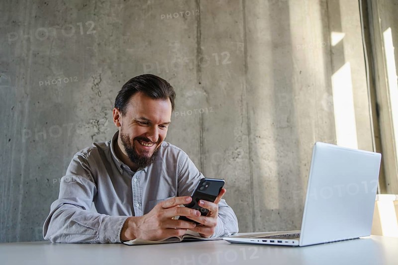 A man in a casual shirt sits at a desk, smiling while using his smartphone. Nearby, a laptop is open, indicating a relaxed yet productive work environment