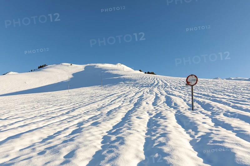 A serene snowy landscape unfolds under a clear blue sky, with a solitary road sign standing amidst the pristine, untouched snow, casting long shadows in the sunlight