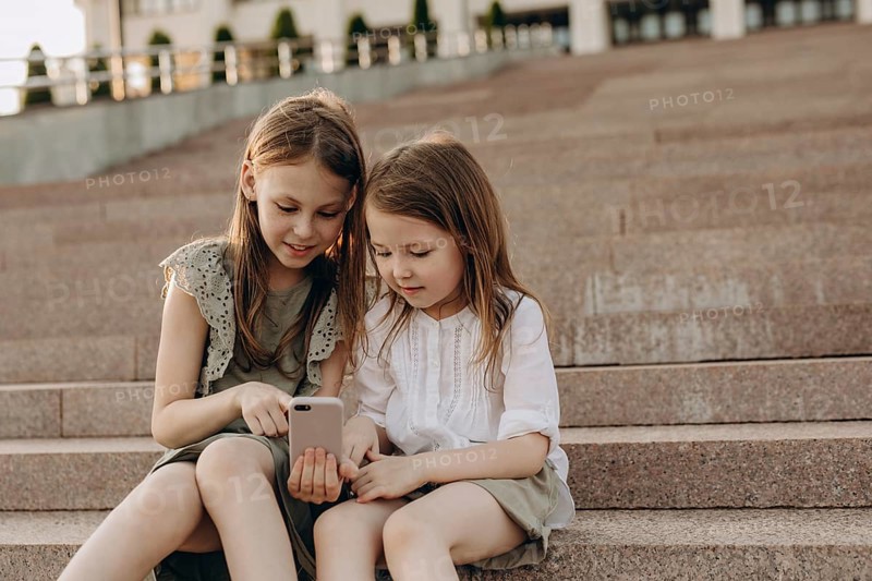 Two young girls sit together on outdoor steps in summer, engrossed in a smartphone. They share a moment of curiosity and exploration, embodying friendship and technology