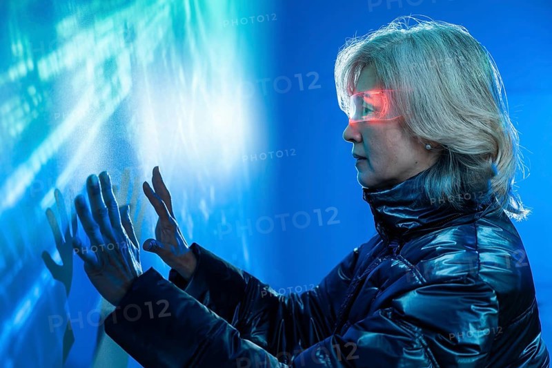 Elderly Asian woman immerses in the metaverse, exploring holographic digital interfaces. Wearing a futuristic jacket, she touches virtual elements, depicting advanced technology use