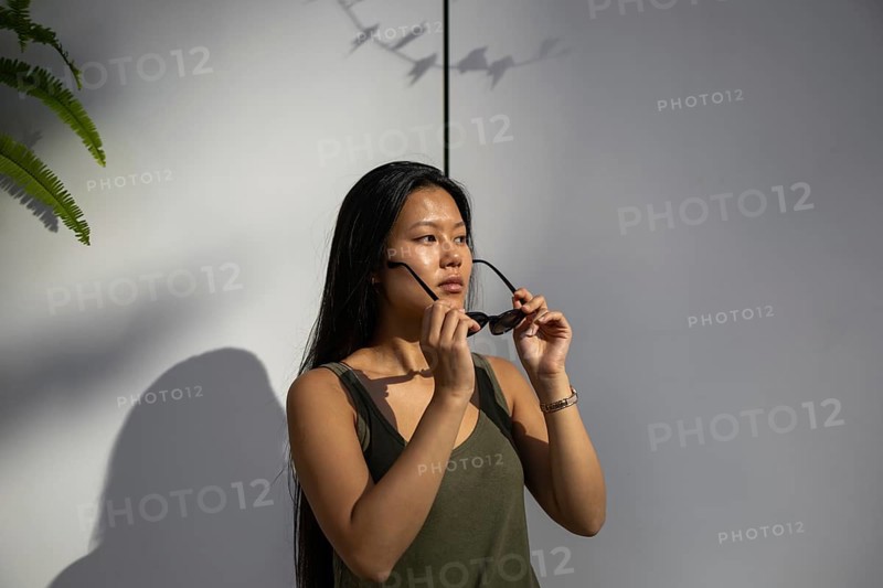 A Chinese woman enjoys a sunny summer day in the city, adjusting her sunglasses while standing against a modern urban backdrop. Her expression conveys relaxation and serenity