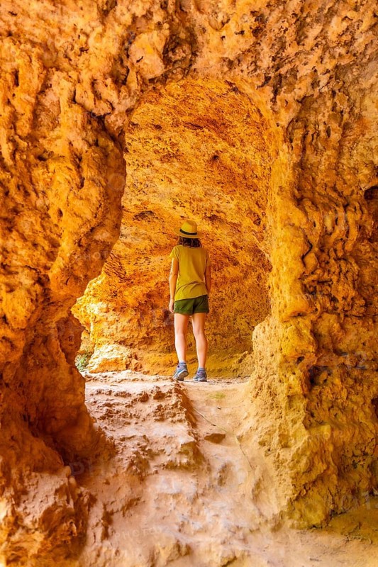 A woman stands in a cave with a yellow rock wall. She is wearing a green hat and shorts