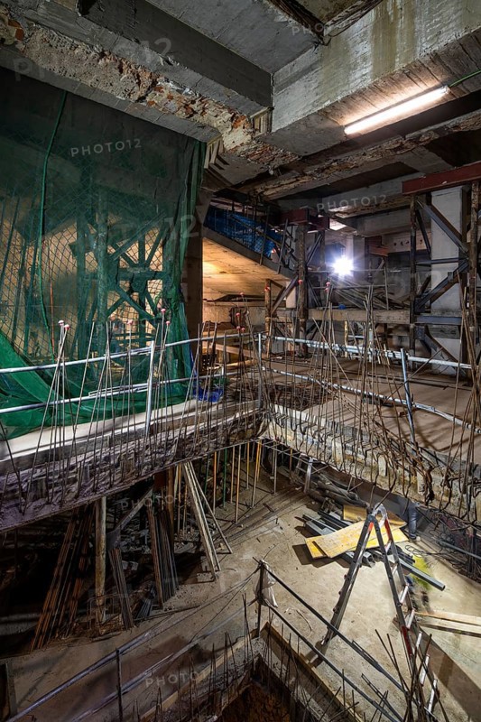 An interior construction site with exposed beams, scaffolding, and safety nets. Illuminated by bright lights, it shows progress and materials in a dynamic work environment