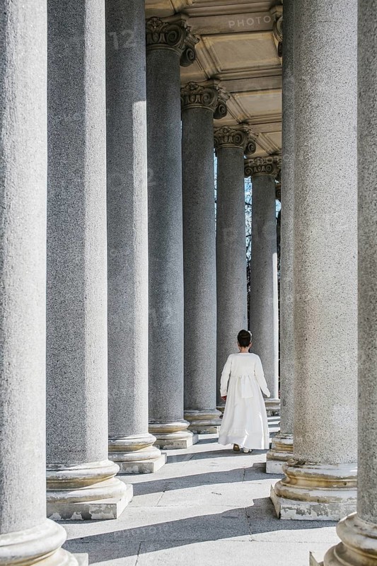 A young girl walks gracefully among towering columns, dressed for her First Communion. The sunlight enhances the serene atmosphere and highlights the elegance of the moment