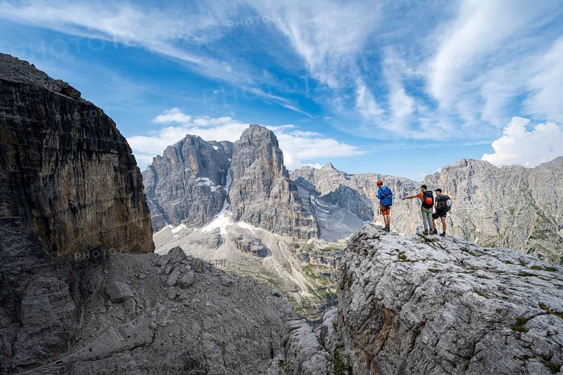 Three mountaineers on a rock in front of a picturesque mountain landscape with rocky peaks, Via Ferrata SOSAT via ferrata, summit of Cima Tosa in the back, Brenta Mountains, Trentino, Italy