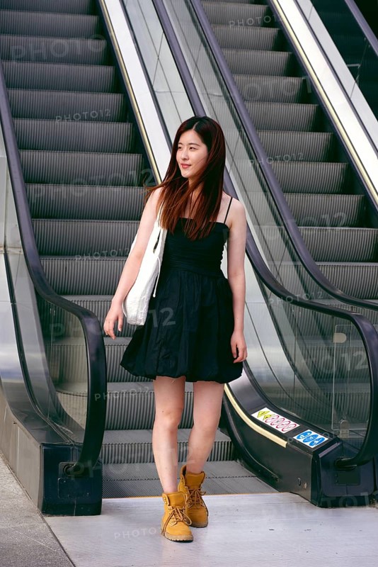 A young asian woman in a black dress stands confidently on an escalator. Her brown hair is loose, and she carries a white bag, highlighting a casual yet stylish urban look