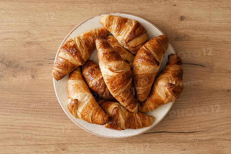 A top view of freshly baked homemade croissants arranged on a white plate, placed on a wooden surface. The golden brown pastries evoke a warm, inviting atmosphere
