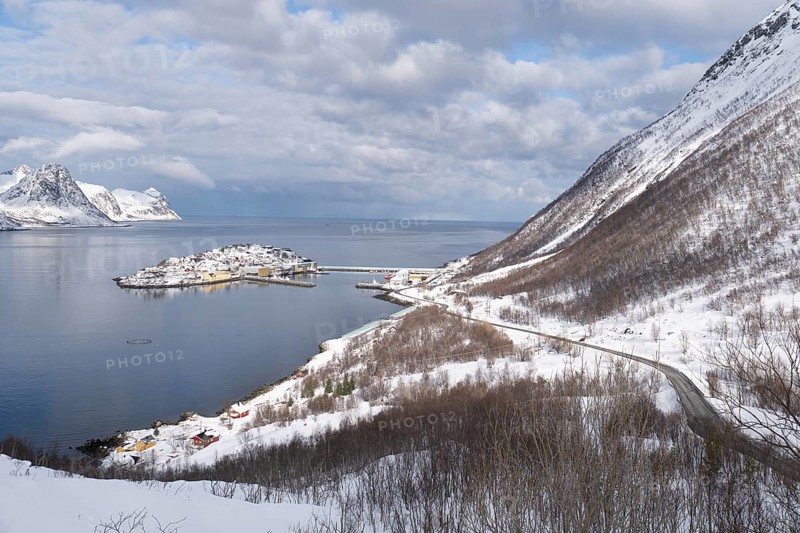 Serene winter view of Husoy, Norway, with a snow-covered coastline, calm sea, and dramatic mountains under a partly cloudy sky. A picturesque northern village in white