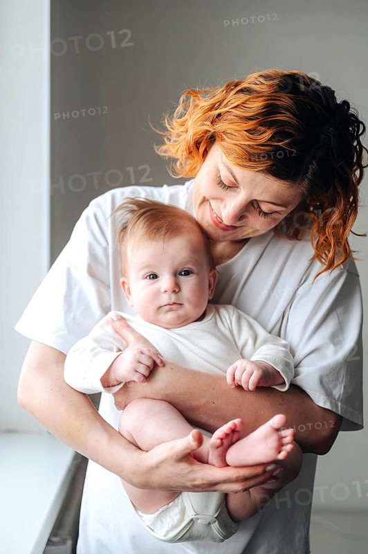 A mother lovingly holds her newborn baby girl, captured in warm natural light. The white clothing enhances the gentle and tender moment shared between them