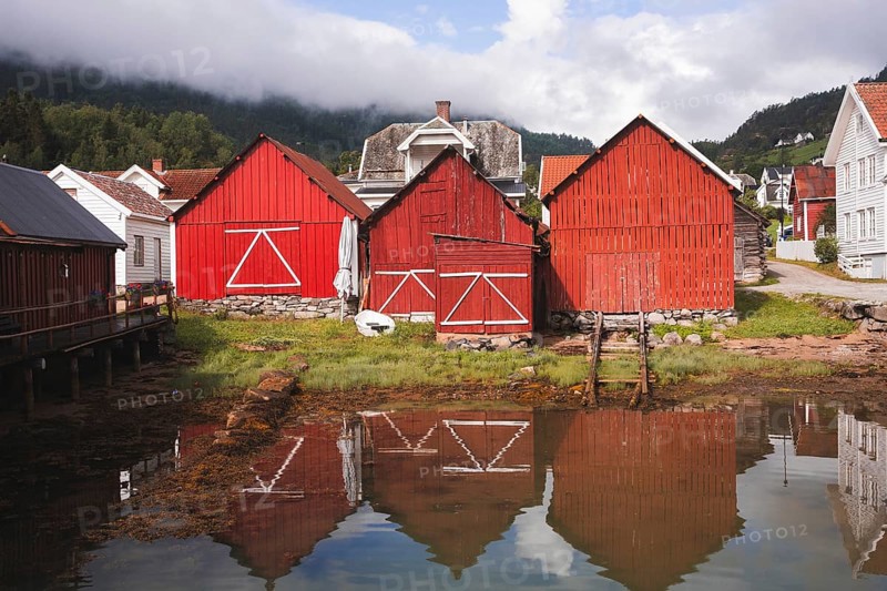 Charming red wooden huts reflect in the calm water of a Norwegian fjord village Surrounded by lush greenery and misty mountains, perfect for outdoor adventures, trekking, or camping
