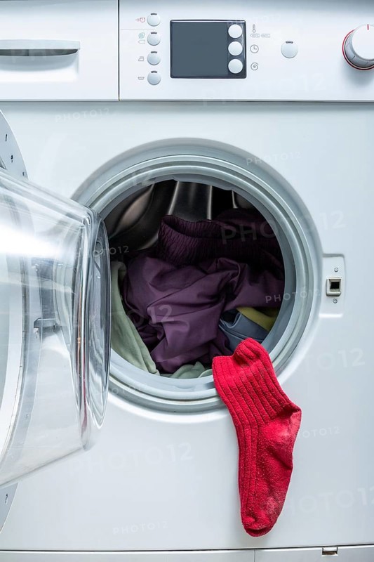 A bright red sock hangs from the door of an open front-load washing machine filled with laundry, emphasizing a humorous and relatable laundry moment at home