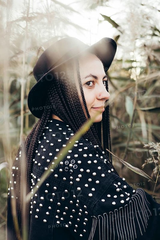 A woman in a unique studded jacket and black hat stands amidst tall grasses at sunset, exuding a chic, mysterious vibe, as the warm light highlights her braided hair