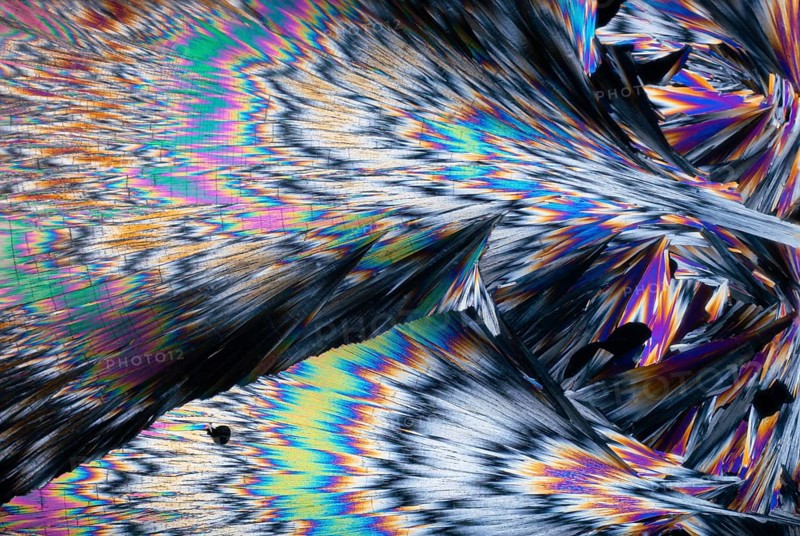Striking, spectrum-like colors of hippuric acid crystals under a microscope. These colors range from vibrant blues, greens, pinks, to fiery oranges and yellows, captured in a dynamic, feather-like formation