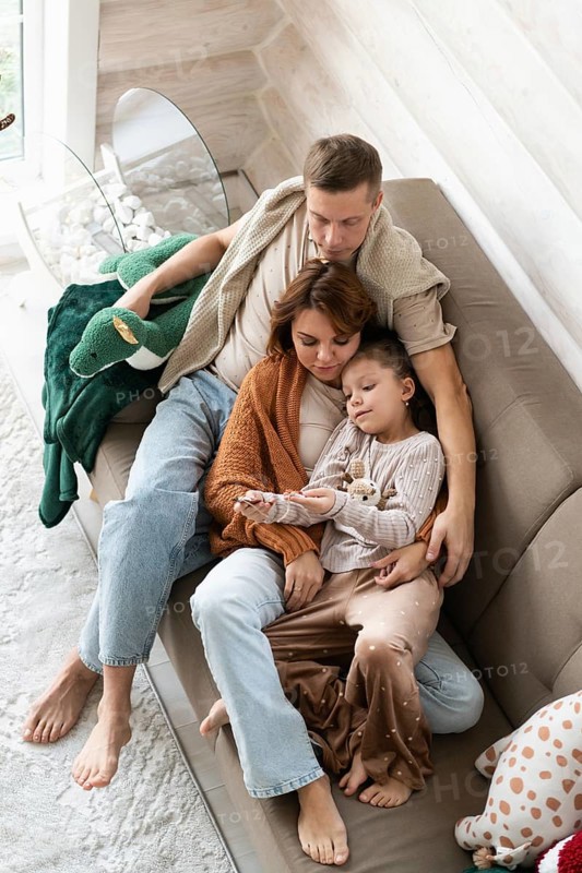 Top view image of a family of four celebrates Christmas in a warm, cozy setting. The mother, holding a child. The father, also seated, gently places his arm around both the mother and the child, in a serene, homely background with festive decorations