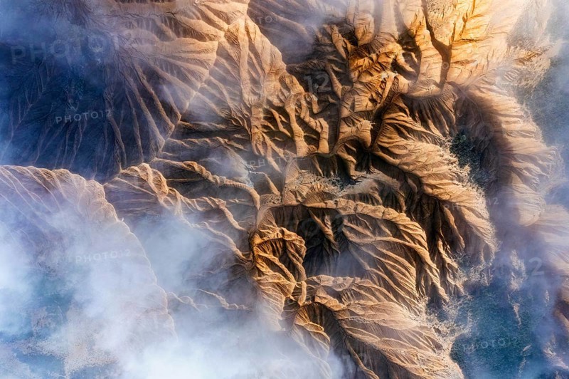 An aerial view showcases rocky mountain formations with deep ridges and valleys Wisps of mist partially obscure the rugged terrain, creating a mystical atmosphere