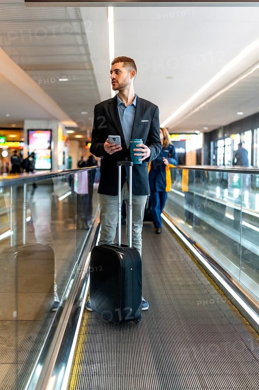 Businessman holding a smartphone and coffee while standing on a moving walkway in an airport terminal