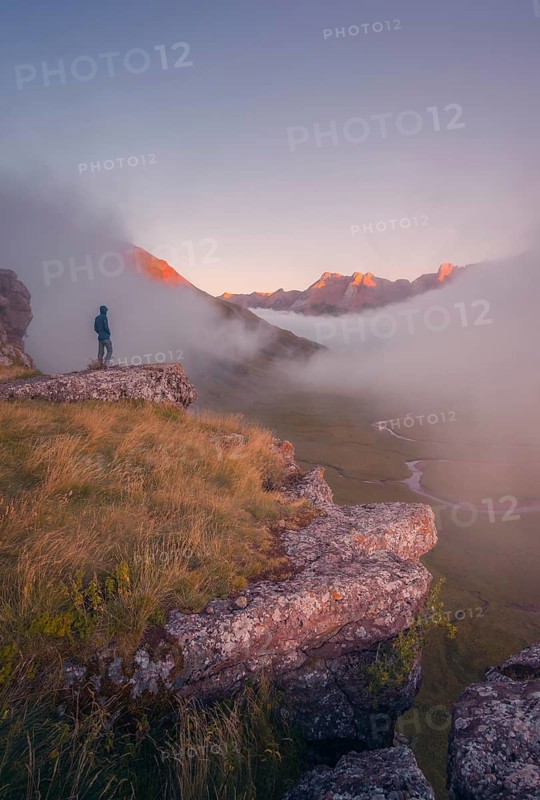 A lone hiker stands on a cliff, gazing at a stunning, mist covered valley at sunrise in the Pyrenees. The early light bathes distant peaks in golden hues, creating a tranquil scene in summer