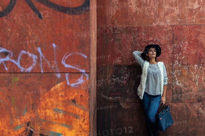 A stylish woman in casual attire with a hat leans against a graffiti-covered wall, showcasing urban fashion. The orange and red background adds a warm, textured contrast