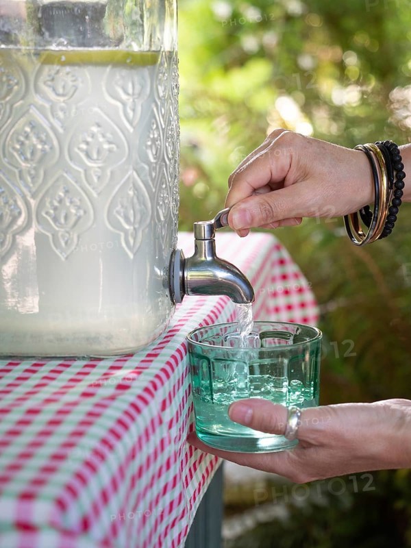 Fresh lemonade in large glass bottle with tap. Hands serving in glass. Refreshing summer drink. Liquid dispenser, sustainable and recyclable containers