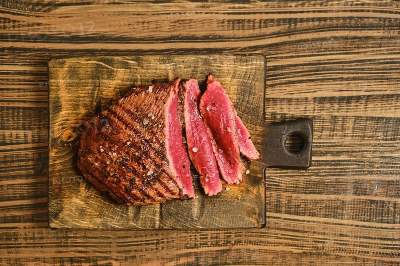 Beef is sliced into thick pieces and placed on a wooden cutting board. The meat shows a reddish color and appears cooked. Seasoning is sprinkled on top. The background is a rustic wooden table