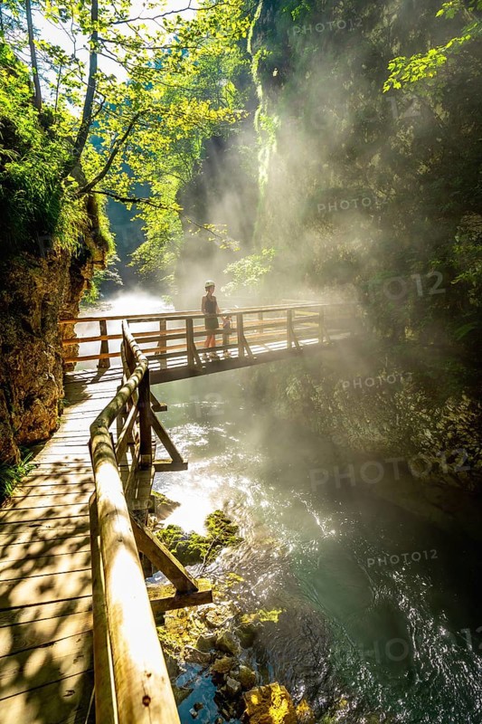 Sunbeams illuminating mist rising from radovna river in vintgar gorge near bled, with tourist walking on wooden footbridge