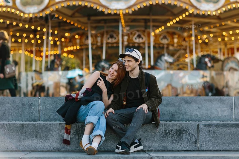 A couple sits on steps, embracing in front of a lit up carousel in Brooklyn Bridge Park. The joyful and warm scene captures a moment of togetherness