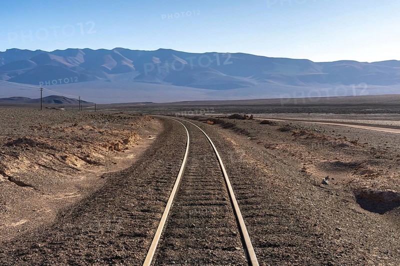 Curving railroad tracks lead through the arid landscape of La Puna, Argentina, with distant mountains under a clear sky