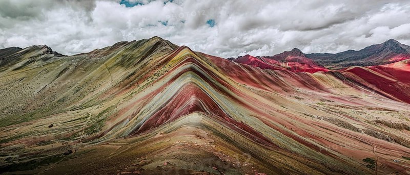 A stunning panoramic capture of Vinicunca, or Rainbow Mountain, famous in Peru for its colorful, layered mineral deposits, under a dramatic sky