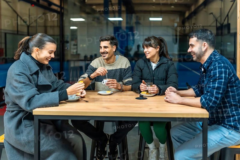Four diverse friends are sitting at a wooden table, drinking coffee and talking, relaxing together after playing pickleball on the court visible through the window