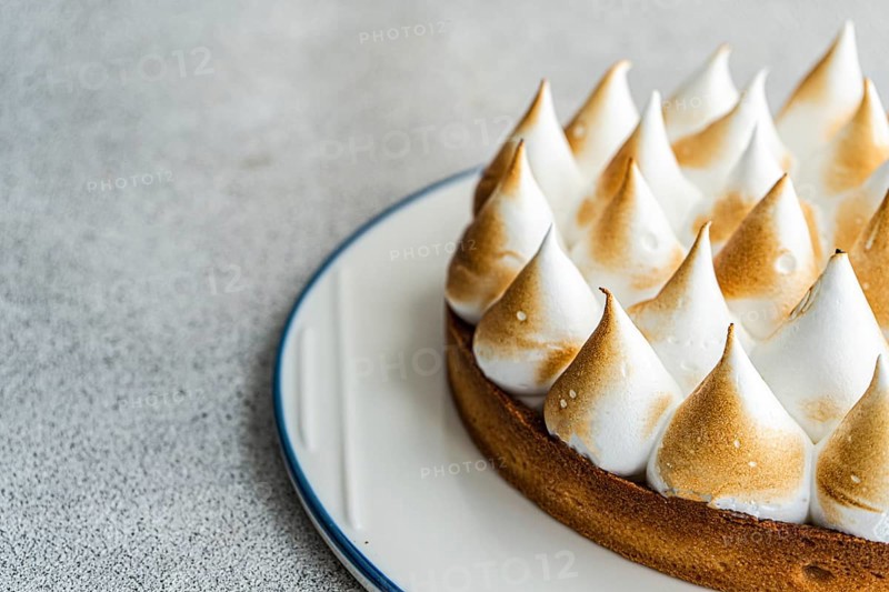 Close up image of a homemade lemon cake, showcasing its golden brown peaks and buttery crust. The dessert is elegantly presented on a white plate against a textured backdrop