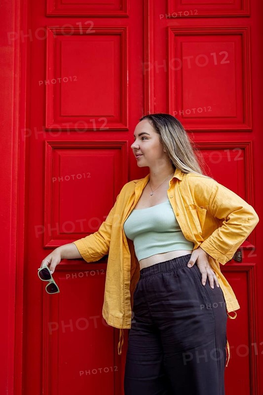 A fashionable woman in a vibrant yellow shirt stands confidently against a bright red door on a city street, embodying the essence of urban exploration and travel