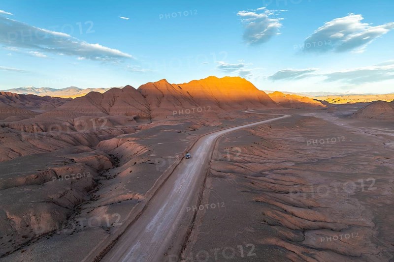 Aerial view of a vehicle in a empty desert road winding through rugged, sunlit mountains at dawn in the argentine Puna. The serene landscape offers a dramatic and peaceful panorama of natural beauty