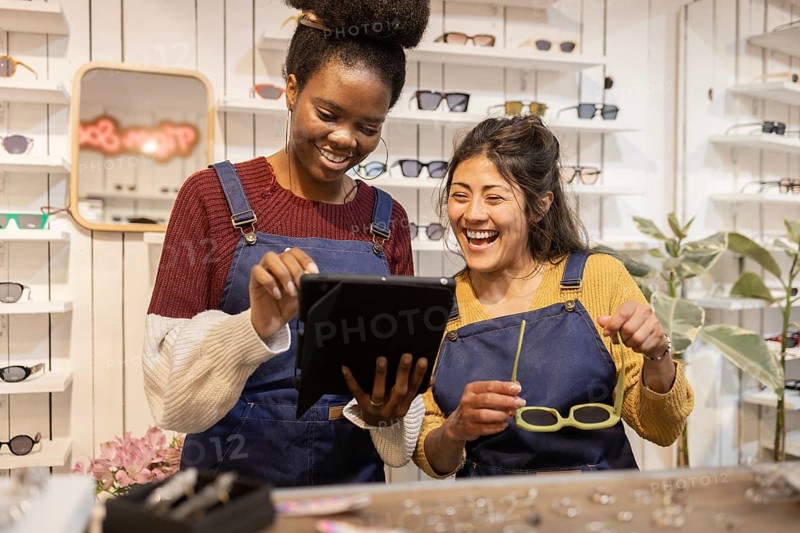 Two cheerful employees in a bustling glasses shop collaborate on a digital tablet, surrounded by a stylish array of eyewear, showcasing teamwork and customer service excellence