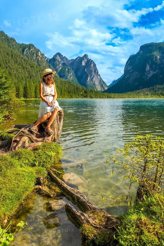 Young woman wearing a straw hat sitting on a tree trunk admiring the landscape of lake dobbiaco in the dolomites