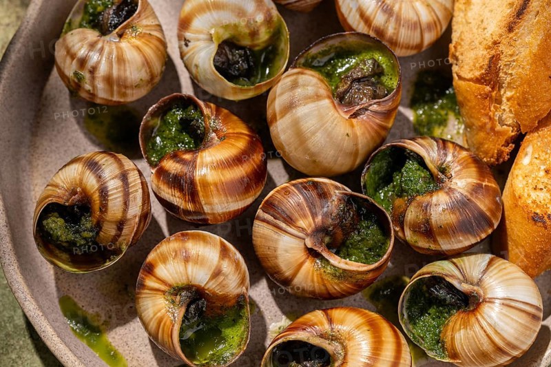 A close-up top view of baked escargot topped with butter and fresh parsley, served alongside crusty bread