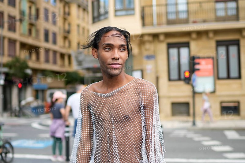 A confident man wearing a mesh top stands on a city street, embodying pride and LGBTQIA+ identity. Urban architecture forms an engaging backdrop
