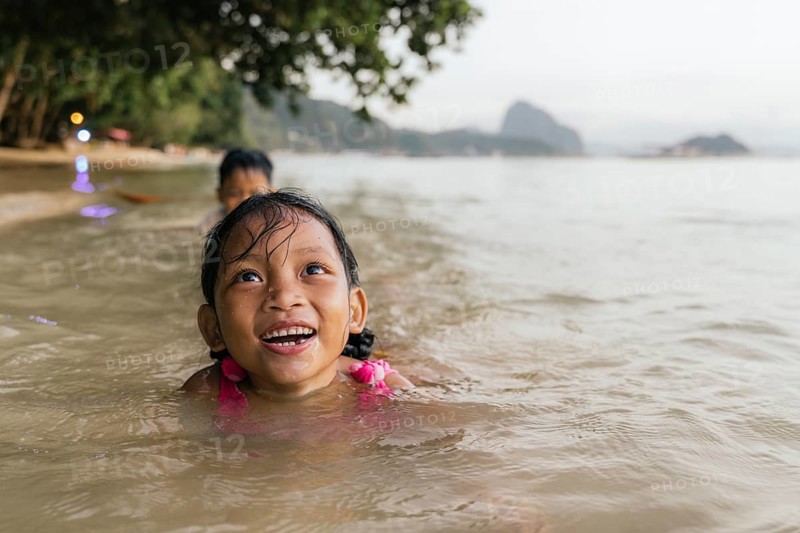 Two children enjoy swimming in the warm, shallow waters of a beach in the Philippines, surrounded by lush green trees, under a clear, bright sky, capturing the joy of carefree playtime
