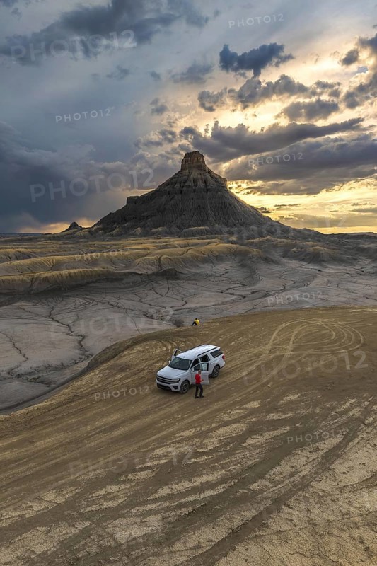 From above an unrecognizable person standing next to a SUV at Factory Butte, Utah, captured during a dramatic sunset with textured cloud-filled skies
