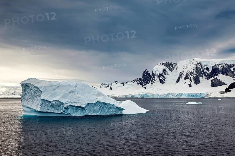 An icy mountain floats in a wide, open sea in front of a dramatic mountain landscape, icebergs in the landscape of the Southern Ocean in Antarctica