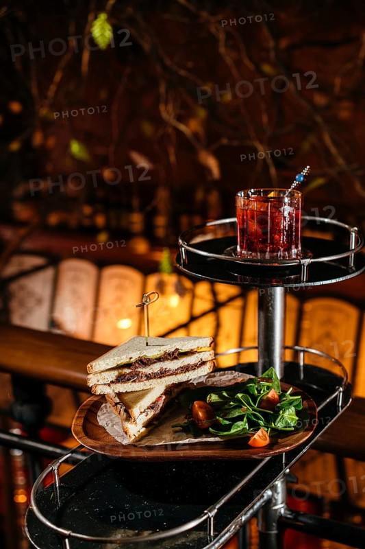 A delectable club sandwich with veal cheeks and cheese, paired with a refreshing Negroni cocktail. Accompanied by a fresh spinach and tomato salad on a wooden table