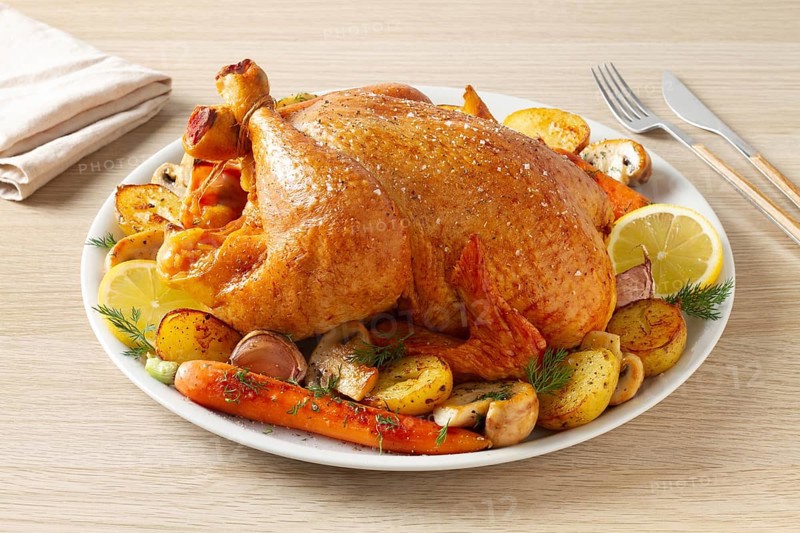 A golden-brown roasted whole chicken is served on a white plate with roasted potatoes, carrots, mushrooms, and lemon slices. A delicious homemade meal perfect for special occasions