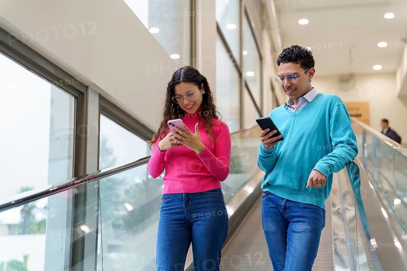 Young man and woman, Gen Z friends, enjoy using their smartphones while riding an escalator in a well-lit modern building, illustrating social media and digital connectivity
