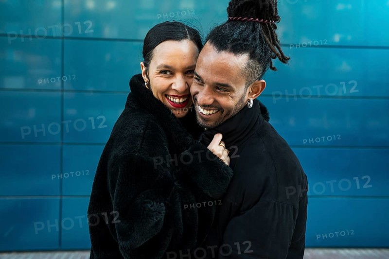 A multicultural couple stands close in a loving embrace, smiling warmly. The urban background adds depth to this candid portrait, highlighting love and diversity