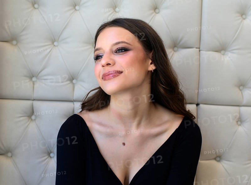 A woman with vibrant makeup and a subtle smile gazes thoughtfully. She is in an elegant setting with a padded wall backdrop, wearing a stylish black top