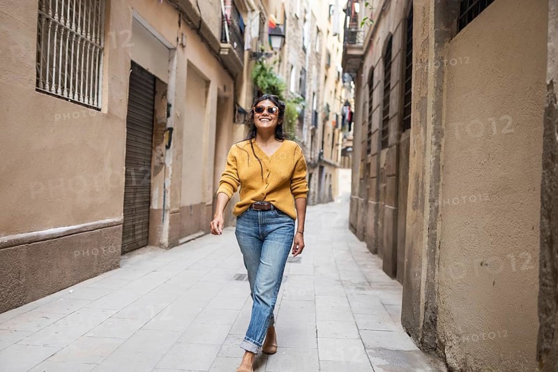 Latin woman in casual attire joyfully walks down a narrow, sunlit city street lined with old buildings. Her confident smile and relaxed demeanor create an inviting atmosphere